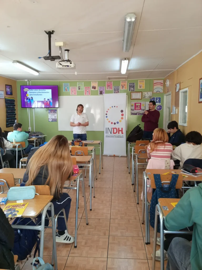 Personas asistiendo a un taller sobre derechos humanos y discriminación en un aula, con un expositor de pie frente a una pizarra y un proyector en el techo. En las paredes hay carteles relacionados con el tema, y un segundo adulto está presente junto a un cartel del INDH.