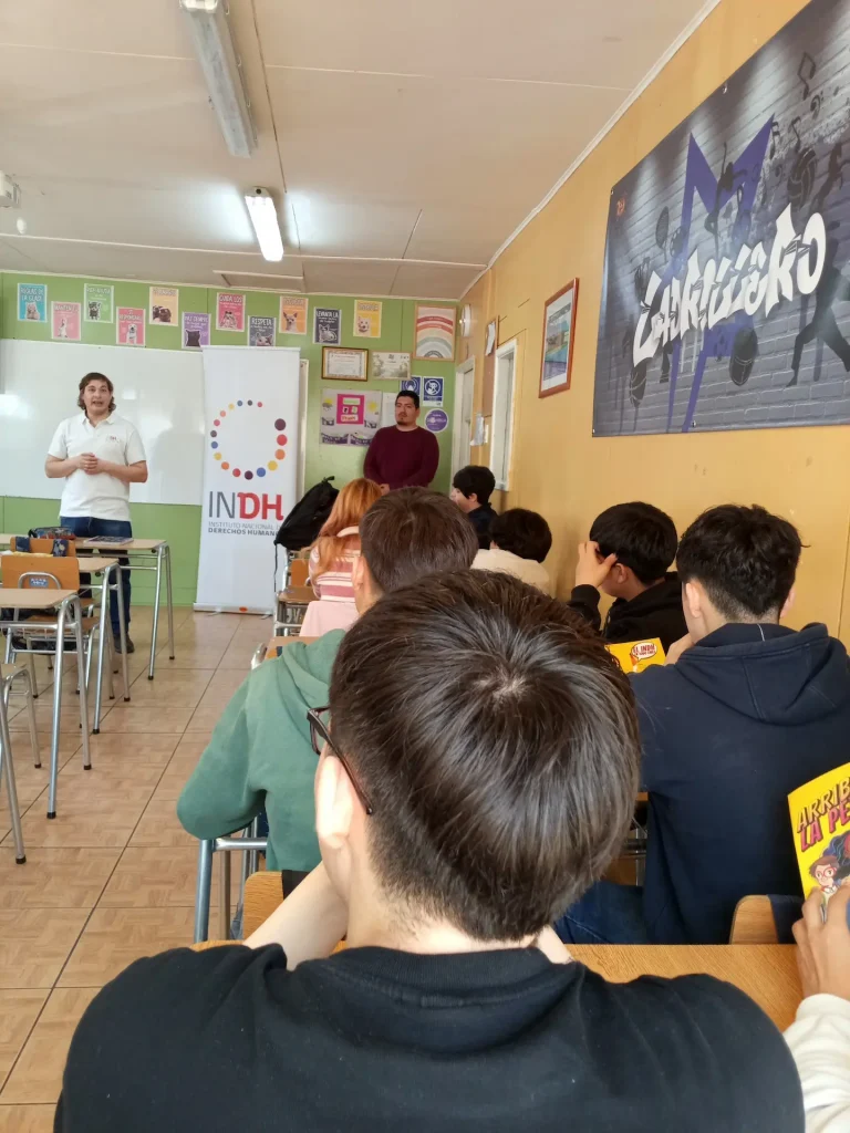 Un aula con estudiantes sentados en escritorios, escuchando a un expositor en el frente junto a un cartel del Instituto Nacional de Derechos Humanos (INDH).