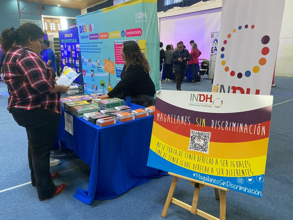 Personas interactuando en un stand del Instituto Nacional de Derechos Humanos (INDH) durante un evento en un gimnasio, con carteles enfocados en la no discriminación en la región de Magallanes.