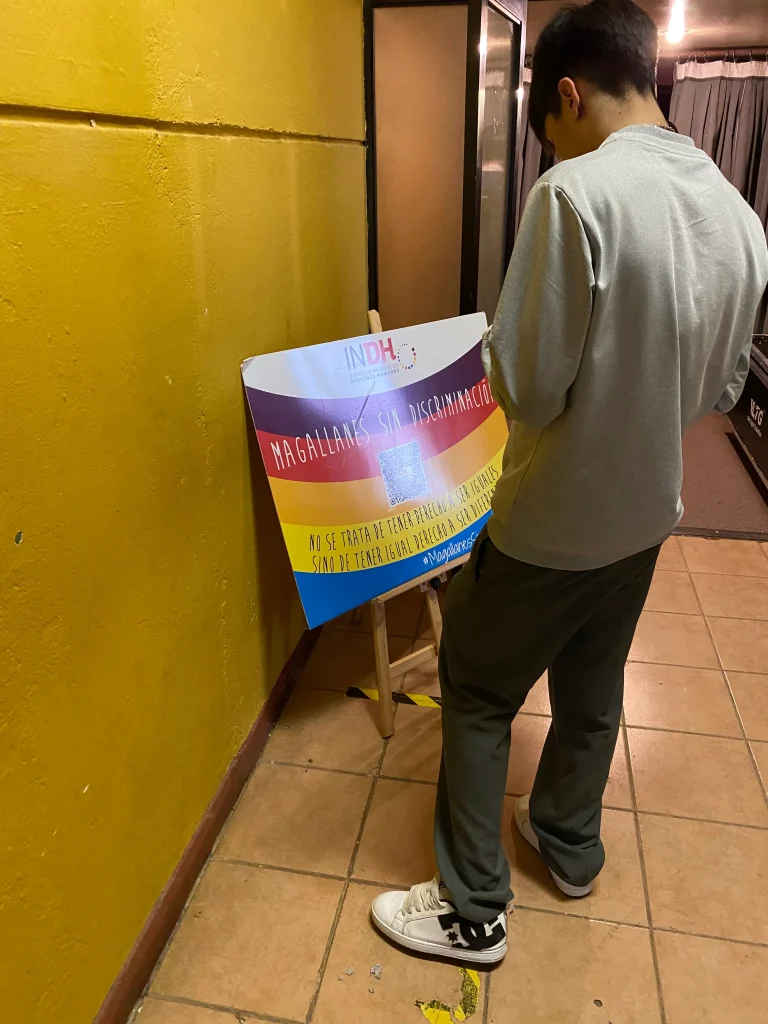 Una persona de espaldas leyendo un cartel con los colores del arcoíris que dice "Magallanes sin discriminación" en un pasillo con paredes amarillas.