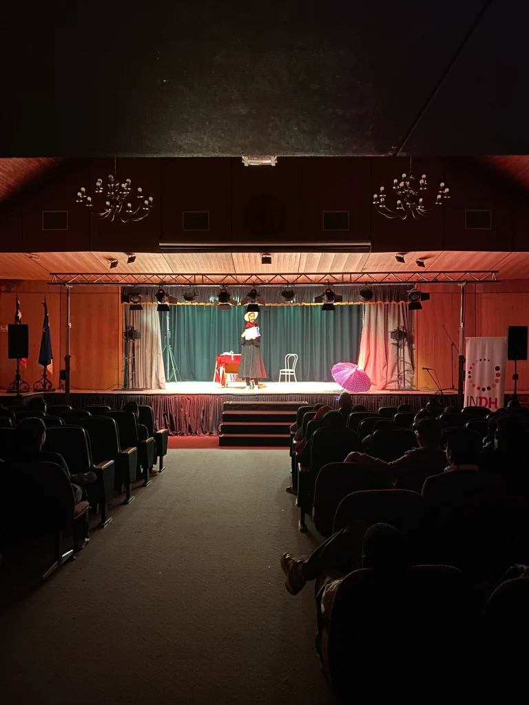 Un artista en el escenario de un teatro iluminado, con un paraguas y una silla a su lado, mientras el público observa desde las butacas en la sombra.