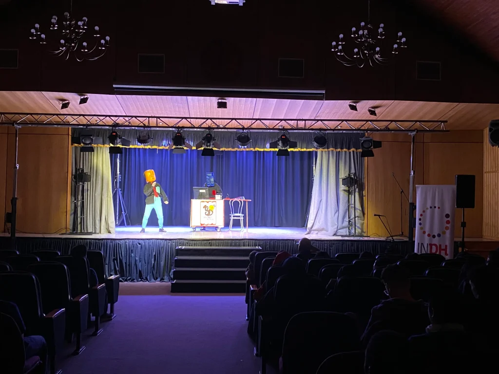 Dos personas con disfraces de cabezas grandes, una naranja y una azul, en un escenario iluminado, actuando frente a un público en un teatro oscuro.