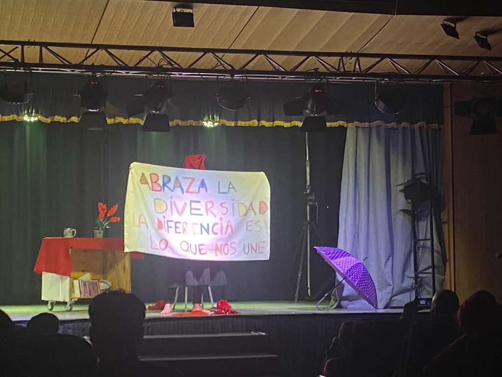 Escenario de teatro con un cartel que dice "Abraza la diversidad, la diferencia es lo que nos une", decorado con una mesa con flores rojas y un paraguas morado.