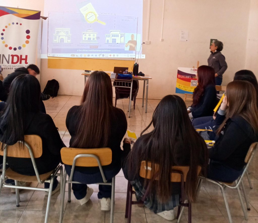 Un grupo de personas sentadas en sillas enfrentan una pantalla de proyección en una sala. En la pantalla aparece una presentación que menciona instituciones como "Ministerios", "Municipios" y "Policías". Hay una persona de pie al frente que parece estar explicando el contenido. A un lado se ve un cartel del Instituto Nacional de Derechos Humanos.