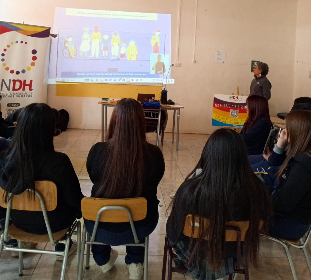 Un grupo de personas sentadas en una sala presenciando una presentación proyectada en la pared. La diapositiva muestra dibujos de personas diversas y un intérprete de lengua de señas. A un lado, hay un cartel y una persona de pie observando. Se aprecia también el logotipo del Instituto Nacional de Derechos Humanos en un cartel cercano.