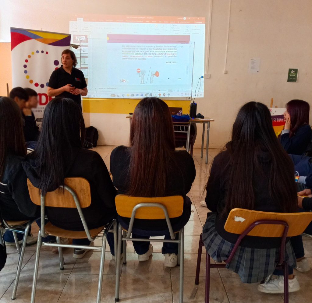 Una persona al frente de un aula hablando a un grupo de estudiantes sentados, con una presentación proyectada en la pared sobre derechos humanos.
