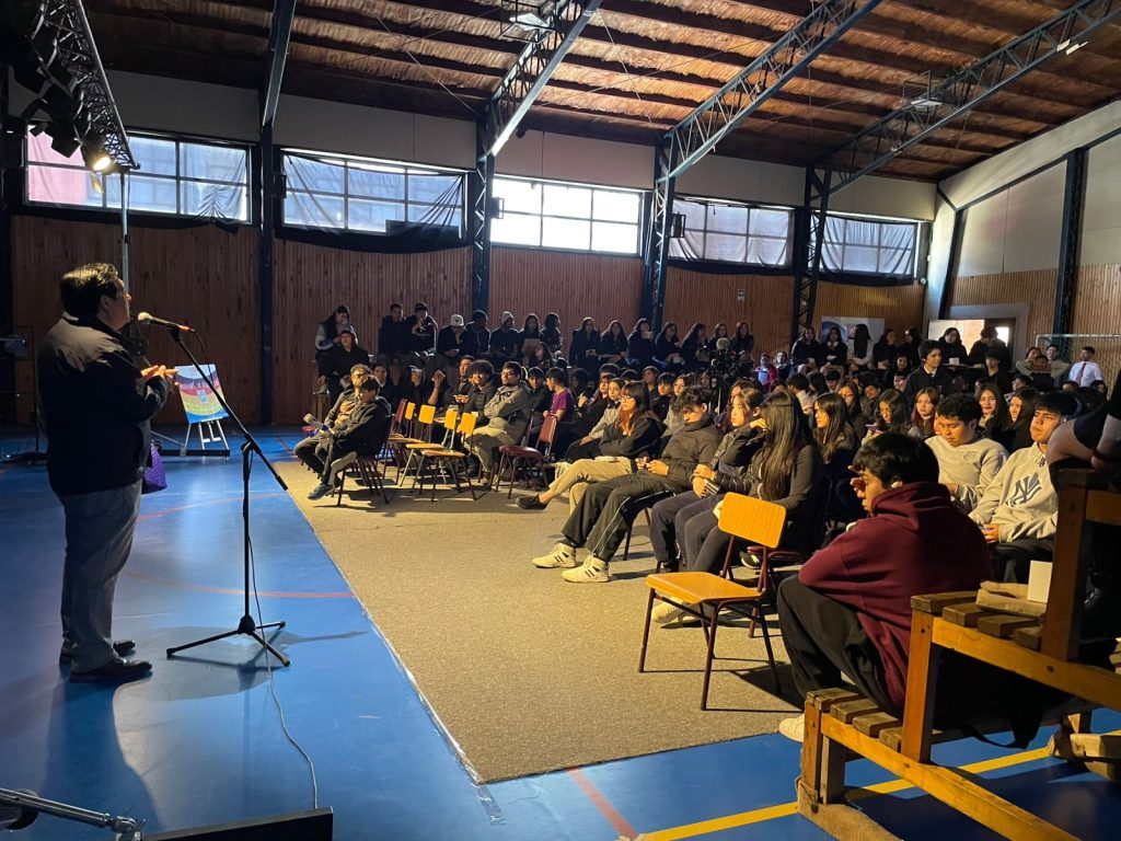 Persona hablando en un micrófono delante de un grupo de personas sentadas en un gimnasio, con el fondo iluminado por luz natural entrando por grandes ventanas.