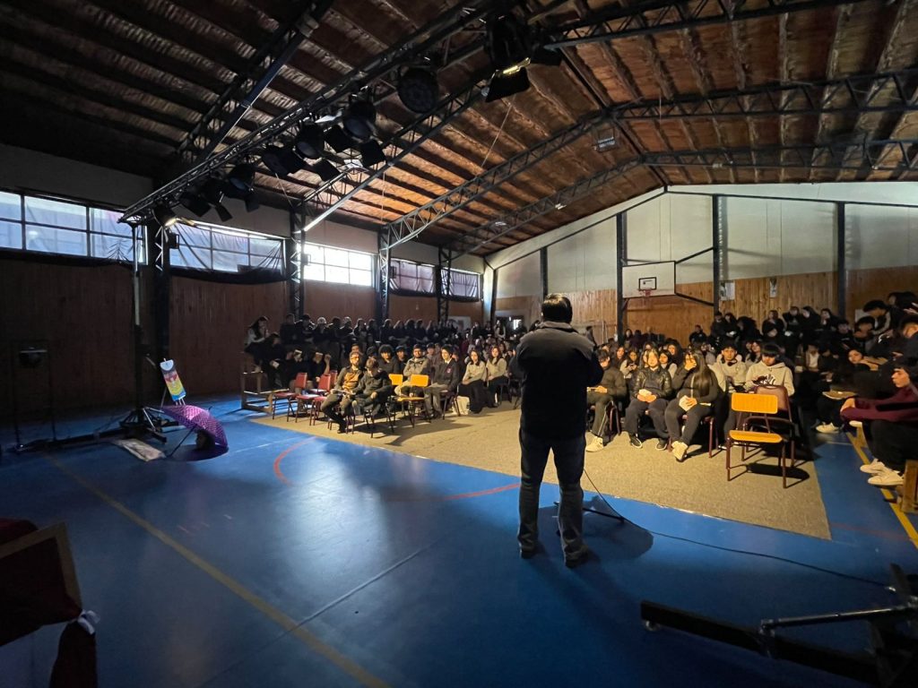 Una persona da una presentación a un grupo de estudiantes sentados en un gimnasio o auditorio, iluminados por focos desde el techo. El área está decorada con una alfombra y posee un ambiente formal con sillas escolares y tribunas.
