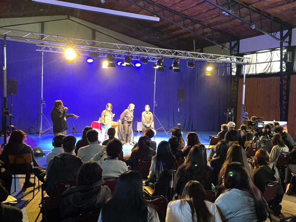 Personas en un escenario iluminado por focos, con una audiencia sentada observando el evento en un recinto cerrado con techo de madera expuesta.