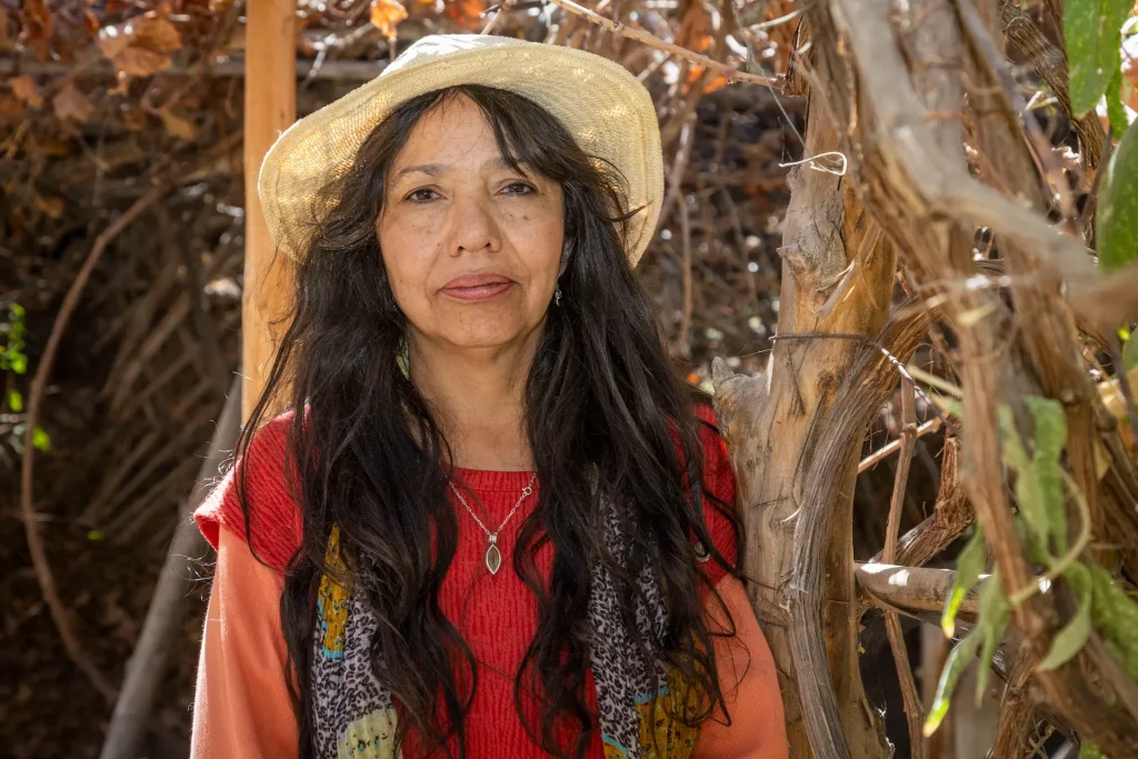 Mujer con sombrero de paja y cabello largo oscuro, vestida con un suéter rojo y bufanda colorida, de pie al lado de un árbol en un entorno natural.
