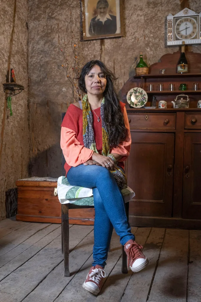 Mujer con cabello largo y bufanda colorida, sentada en una silla sobre un suelo de madera, con un aparador de madera en el fondo y decoraciones antiguas.