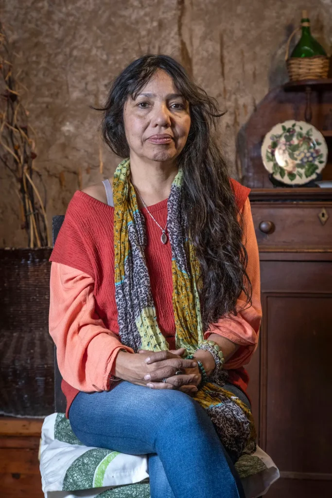 Mujer sentada con cabello largo suelto, lleva un suéter rojo, bufanda colorida y jeans, en un entorno interior con muebles antiguos y decoraciones.