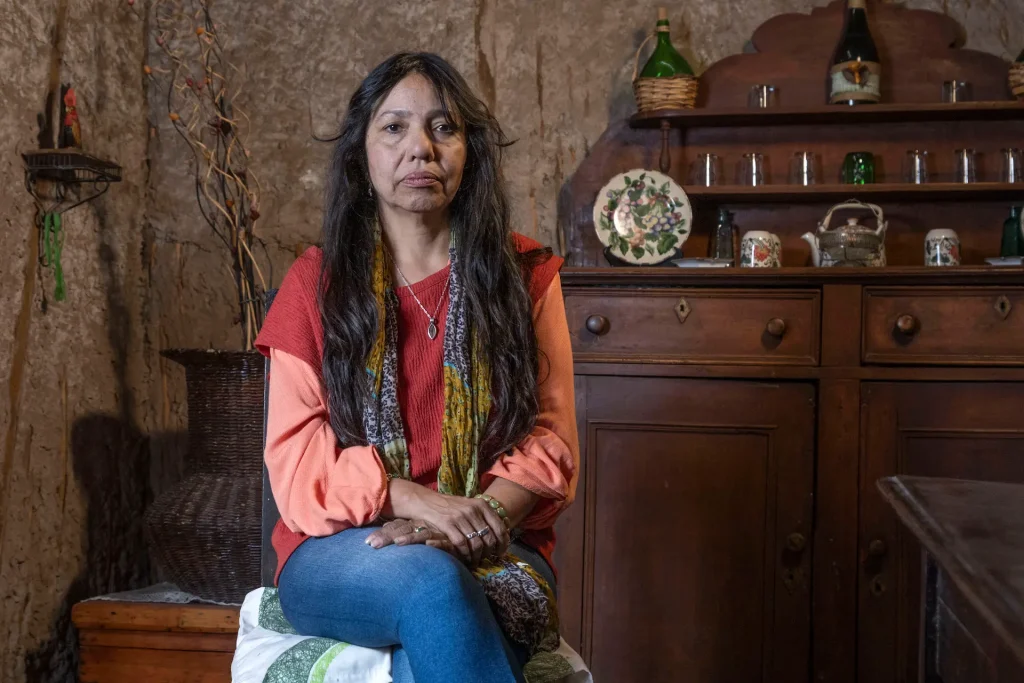 Mujer sentada con las manos cruzadas sobre sus piernas, en una habitación con una pared de piedra y un aparador de madera decorado con tazas, platos y botellas.