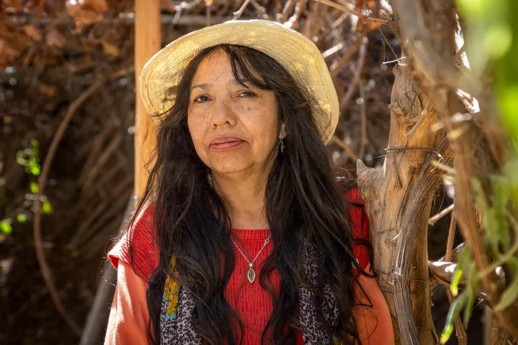 Mujer de cabello largo y sombrero, vestida con suéter rojo, posa entre ramas secas en un entorno natural.
