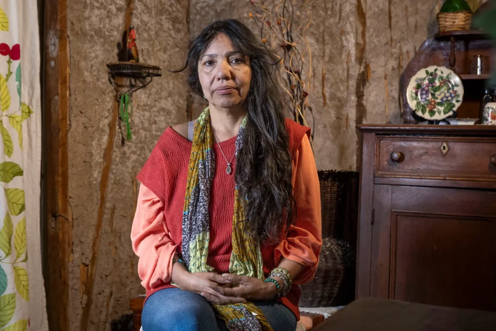 Retrato de Patricia Zúñiga Mitchell. Mujer sentada en una habitación rústica con muro de barro, vistiendo un jersey rojo, pantalones de mezclilla y una bufanda colorida; observa hacia la cámara con una expresión serena, y detrás hay un mueble de madera con platos decorativos.