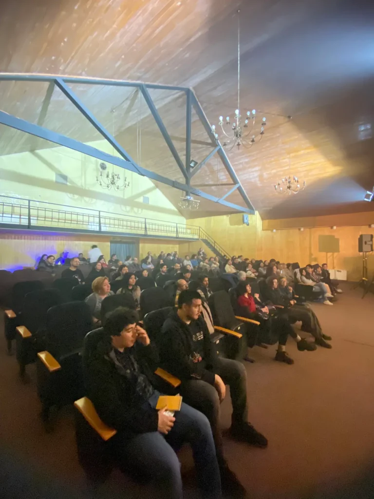 Personas sentadas en una sala de cine o auditorio, observando atentamente hacia el frente; el lugar cuenta con un techo elevado con estructuras metálicas y lámparas de araña.