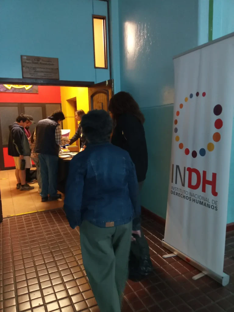 Personas que ingresan a una sala donde hay una mesa con individuos atendiendo detrás, con un cartel del Instituto Nacional de Derechos Humanos al lado de la puerta.