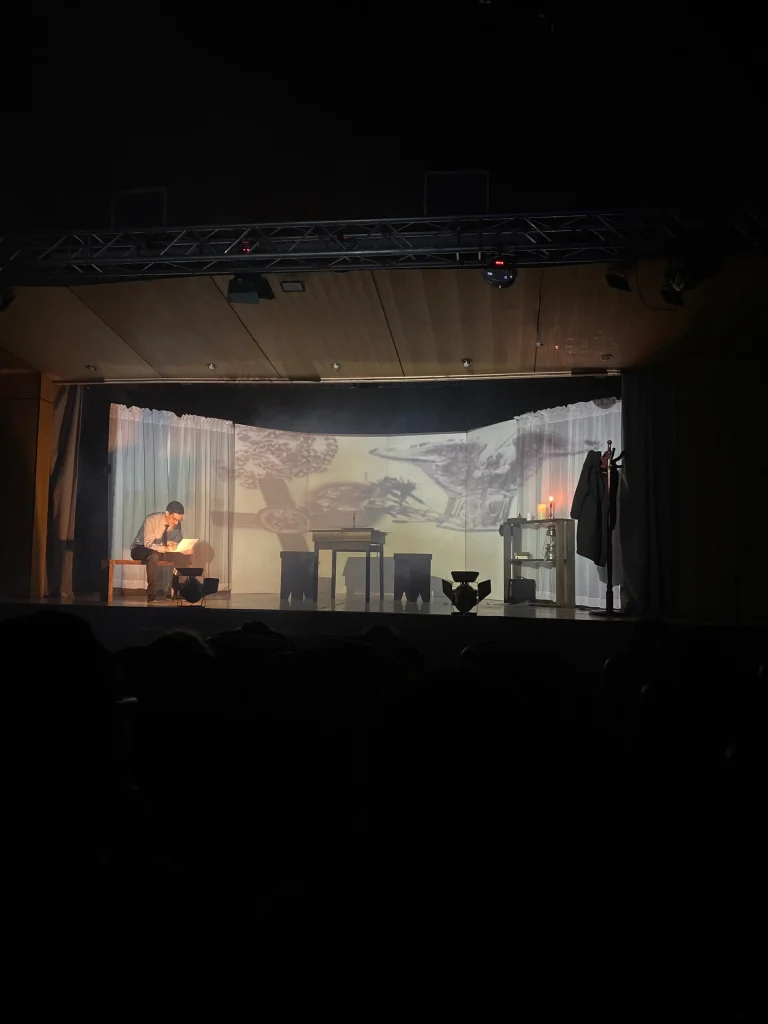 Un escenario con una persona sentada a la izquierda, sosteniendo un papel, y una mesa al centro con una luz tenue. Detrás hay una pantalla con proyecciones artísticas y cortinas a los lados. La iluminación es baja, creando un ambiente dramático.