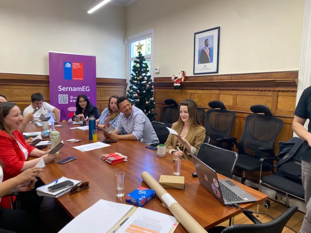 Personas sentadas alrededor de una mesa en una reunión, con carteles de SernamEG y un árbol de Navidad decorado al fondo.