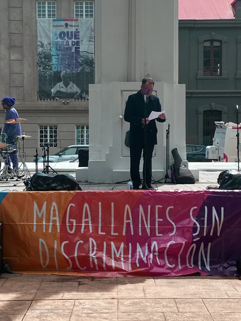 Hombre hablando en un micrófono en un evento al aire libre con una pancarta que dice "Magallanes sin Discriminación".