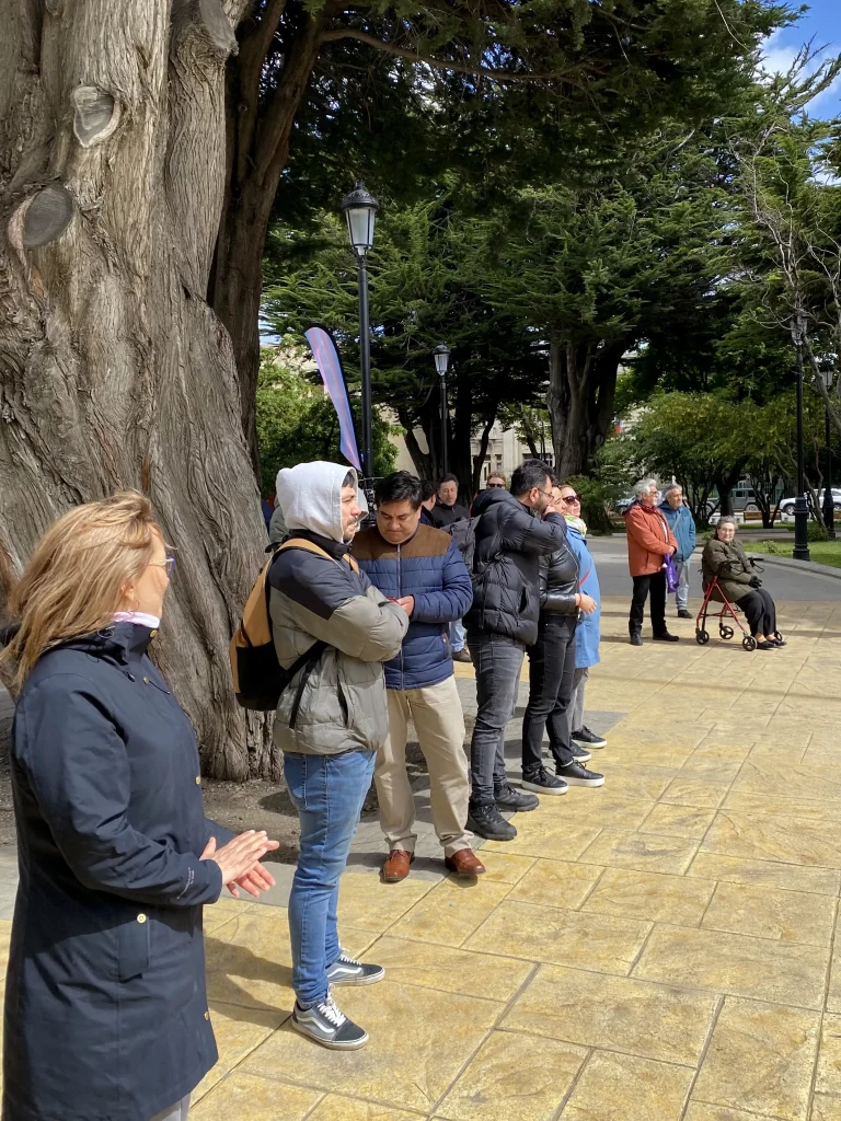 Grupo de personas en un parque, algunas abrigadas, junto a un árbol grande y un camino empedrado.