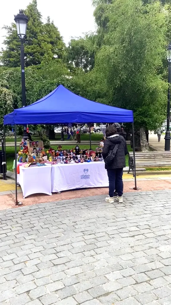 Persona observando una mesa con muñecos artesanales expuesta bajo una carpa azul en un parque.