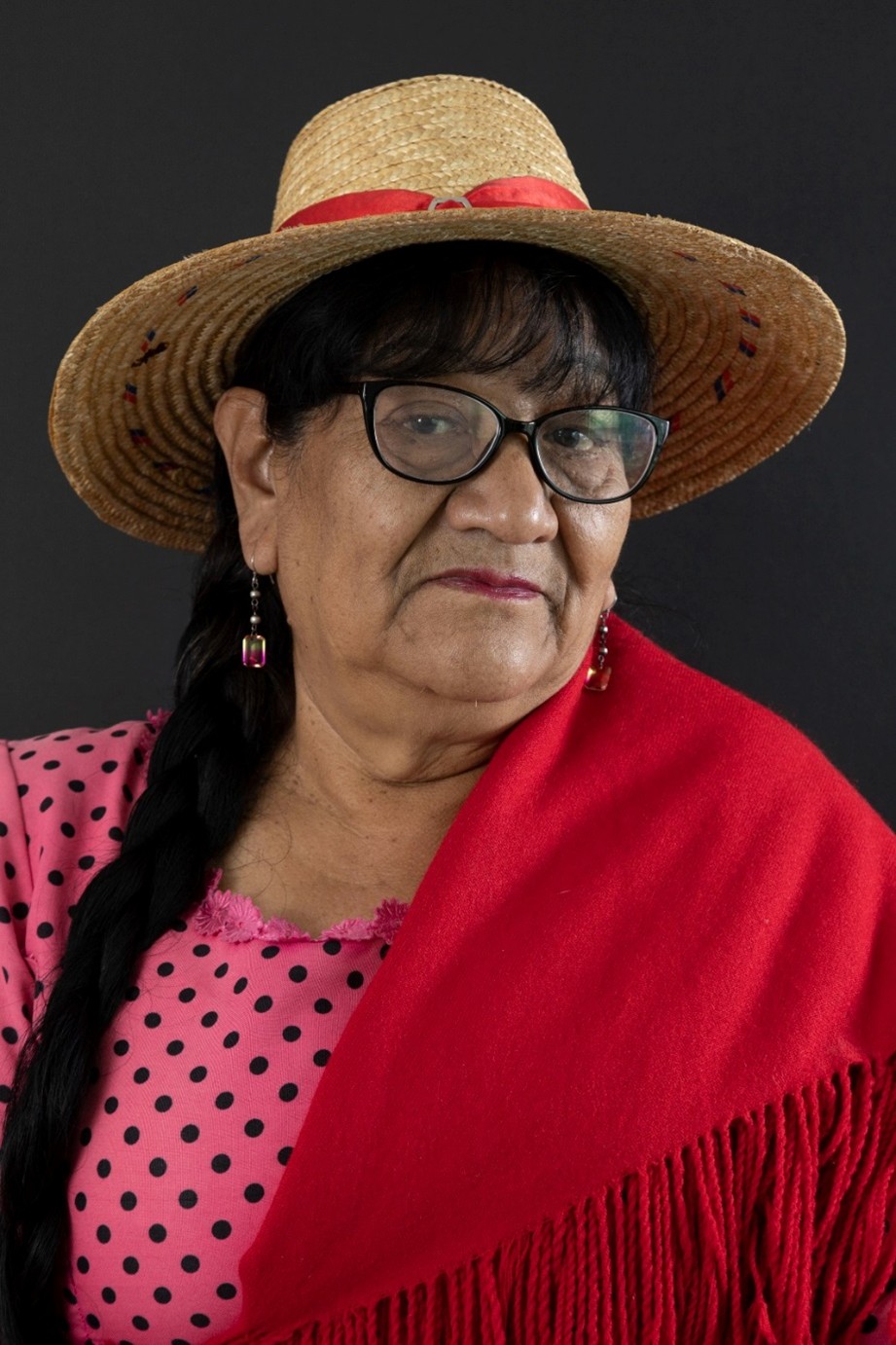 Mujer con gafas, trenza, sombrero de paja, blusa rosa de lunares y chal rojo, en fondo oscuro.