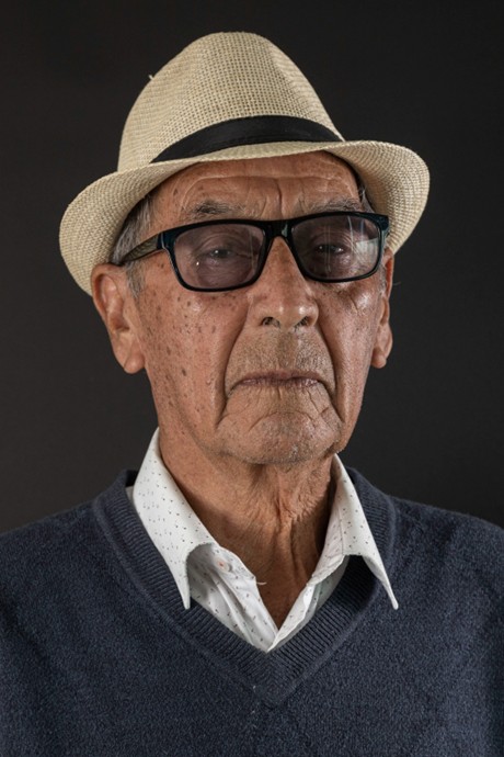 Un hombre mayor con gafas y sombrero de paja, vistiendo un suéter gris y camisa blanca.