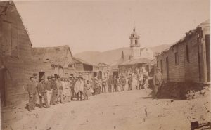 Personas vestidas con ropa de época se alinean en una calle de tierra, con edificios de madera y una iglesia al fondo.