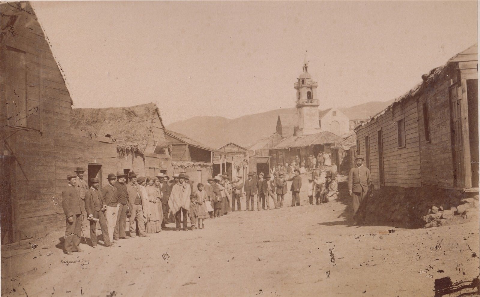 Personas vestidas con ropa de época se alinean en una calle de tierra, con edificios de madera y una iglesia al fondo.
